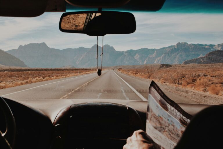 View from inside a car driving down an empty desert highway toward distant mountains, with a person in the passenger seat holding a map